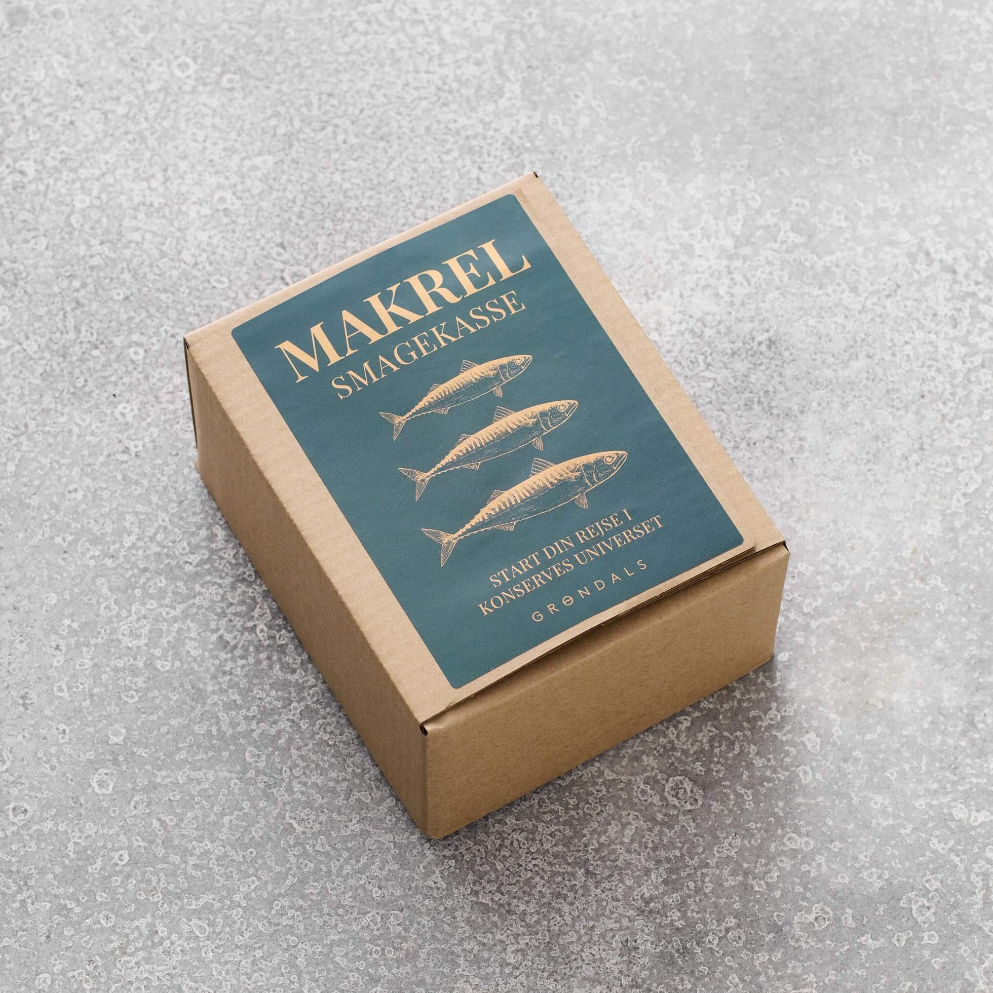 En brun Grøndals Makrel Smagekasse med en blå etiket med tre fiskeillustrationer og dansk tekst, der indeholder gourmetmakrel, står på en lysegrå struktureret overflade.