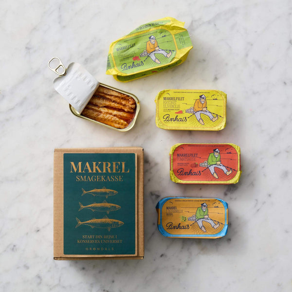 Fire farverige dåser med Grøndals Makrel Smagekasse, inklusive en åbnet dåse med håndfileterede makreller, er pænt arrangeret på en hvid marmoroverflade.