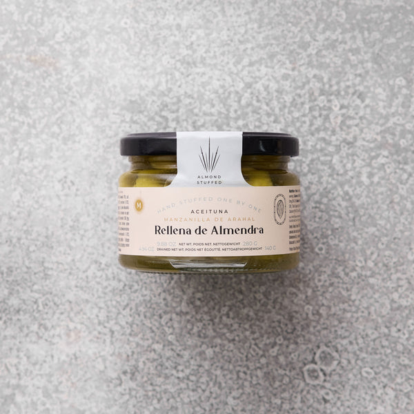 Glaskrukke med Rellena de Almendra, mandelstuffede oliven fra Grøndals, med en beige etiket, set mod en grå tekstureret baggrund.