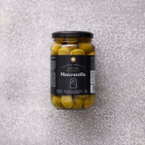 En glaskrukke med Grøndals Manzanilla oliven med sten, med sort låg og etiket, står på en struktureret grå overflade.