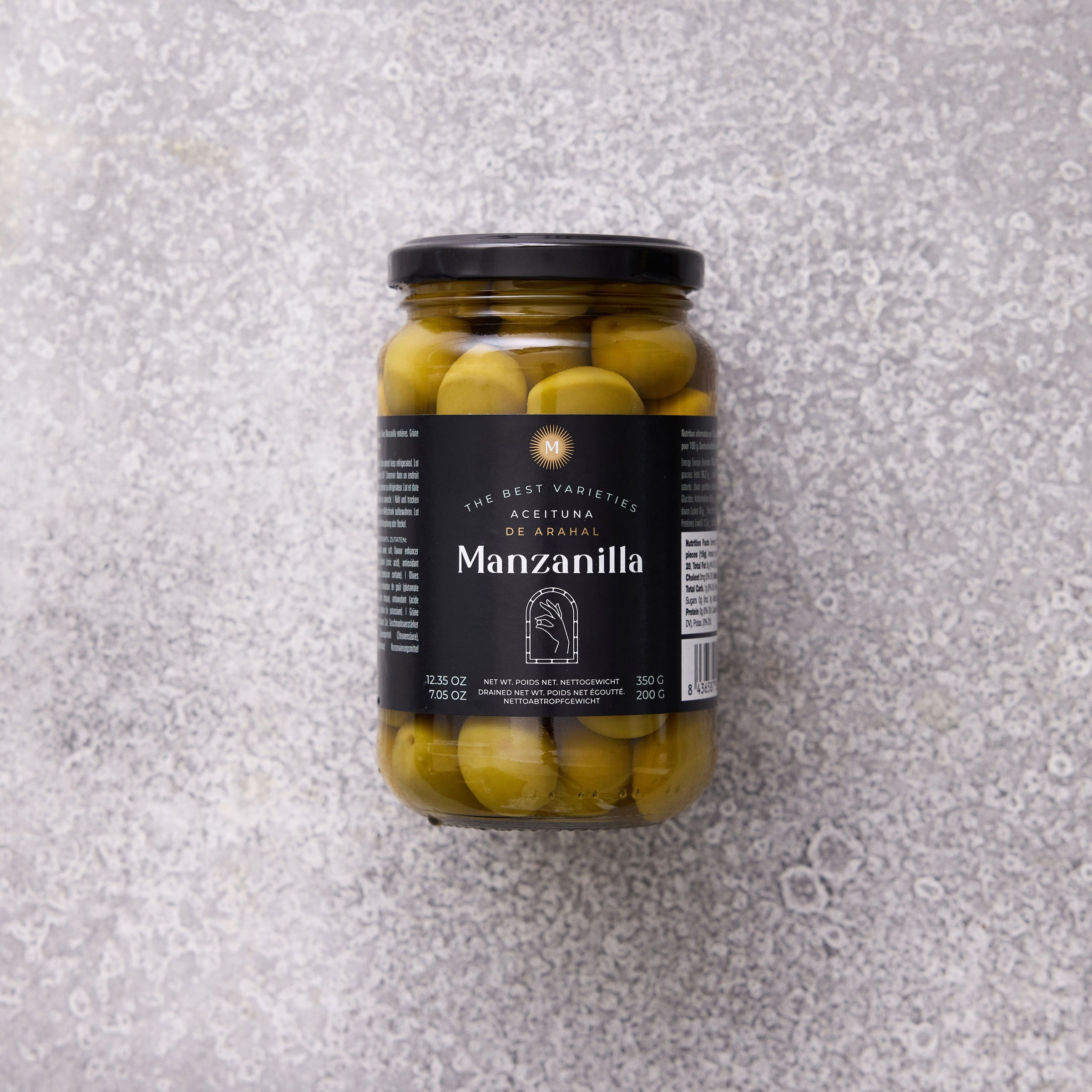 En glaskrukke med Grøndals Manzanilla oliven med sten, med sort låg og etiket, står på en struktureret grå overflade.