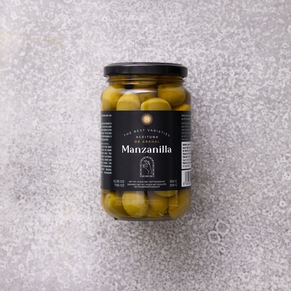 Glaskrukke med Manzanilla oliven fra Grøndals, med en sort etiket, set mod en grå tekstureret baggrund.