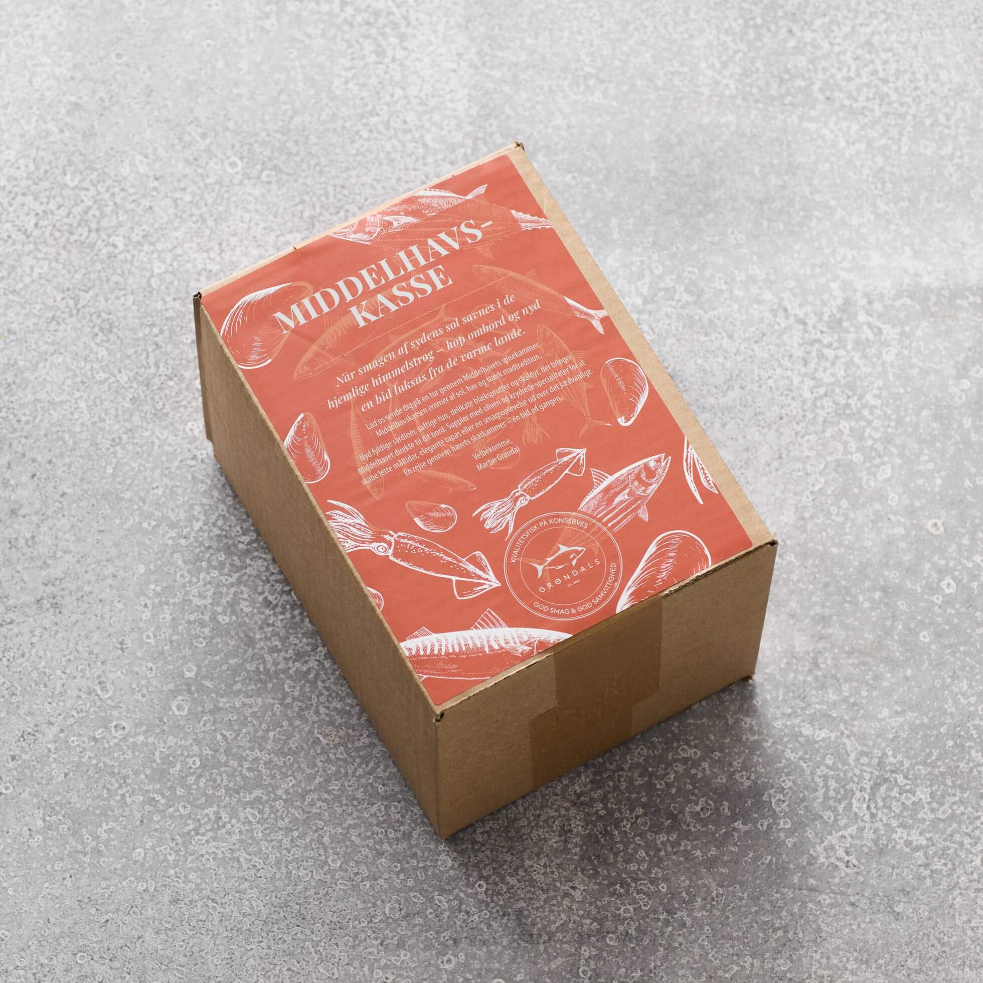 En lukket papkasse mærket Middelhavskassen fra Grøndals hviler på en grå tekstureret overflade, dekoreret med hvide illustrationer af sardiner, rejer og andre skaldyr – ideel for tapasentusiaster.