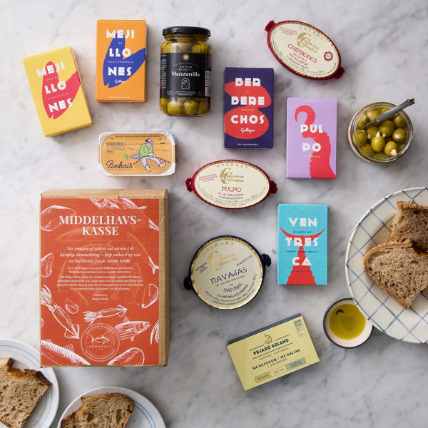Grøndals’ Middelhavskassen byder på middelhavskonserverede fisk og skaldyr, herunder sardiner, samt oliven og brød – smukt arrangeret med farverig emballage og en glasskål med grønne oliven til den perfekte tapas-anretning.
