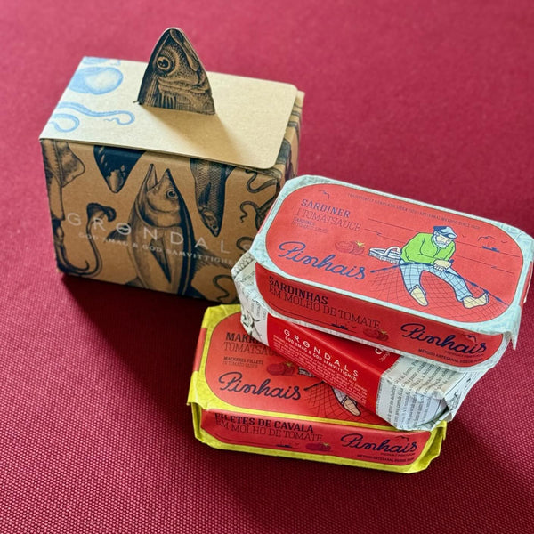 Tre farverige dåser med Grøndals fiskekonserves, stablet på en rød flade, står ved siden af den dekorative "Mors dags kube"-æske med fiskeillustrationer og et håndtag formet som en fiskehale.