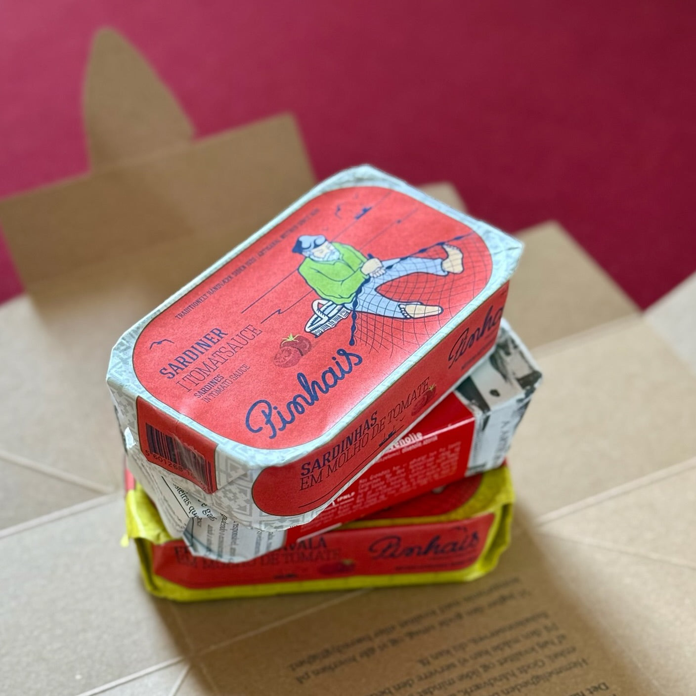 Tre farverige dåser med sardiner, stablet på en åben kasse. Den øverste dåse har en illustration af en person ved vandet med et fiskenet og viser Grøndals-mærket og "Mors dags kube" på etiketten.