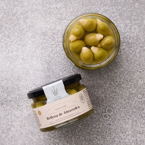 To glaskrukker med Grøndals Manzanilla oliven med mandel står på en grå stenoverflade - den ene er åben, så man kan se de mandelfyldte oliven indeni, som er ideelle til spansk tapas. Den forseglede krukke har etiketten "Rellena de Almendra.