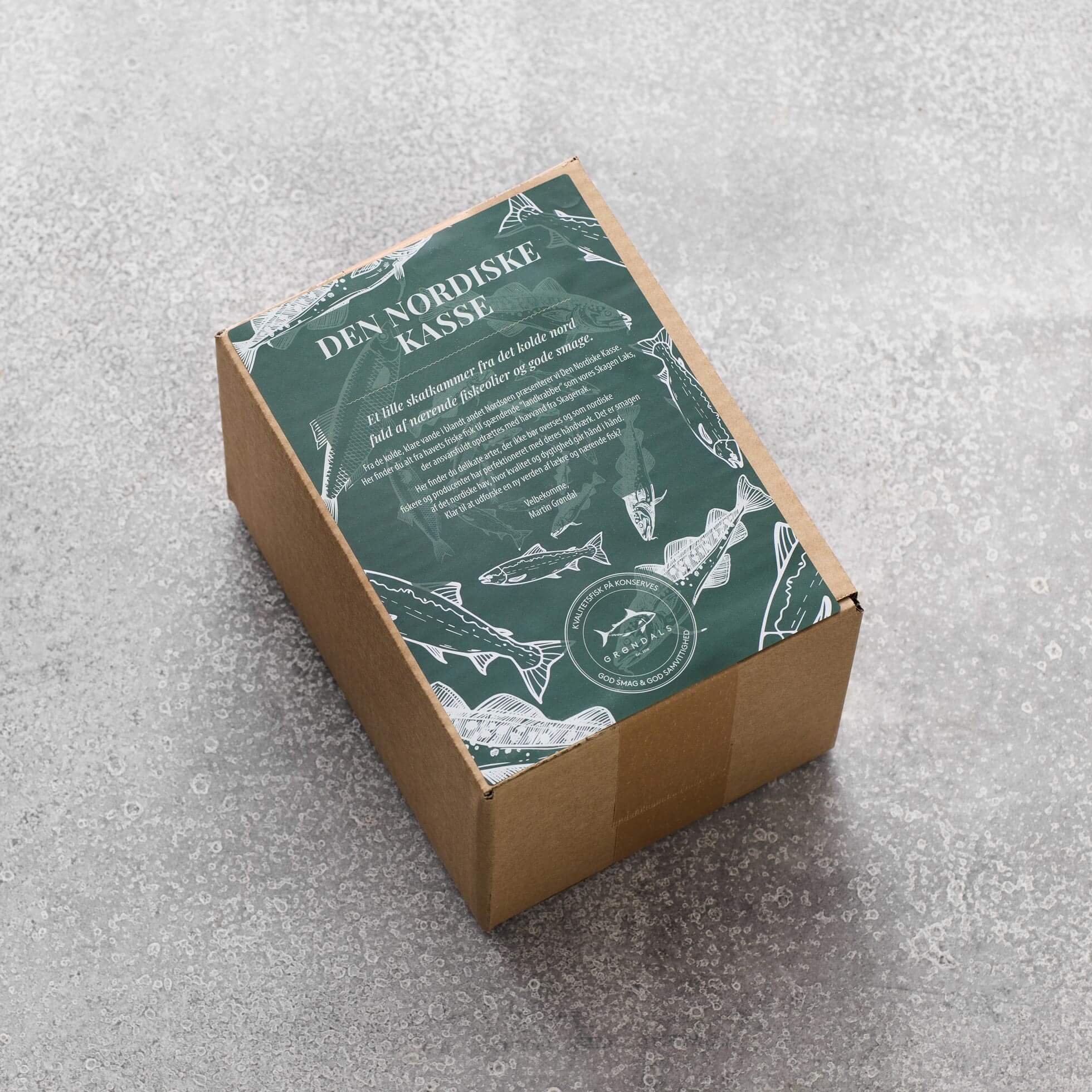 En Grøndals Den Nordiske Kasse-papkasse med en grøn flyer med hvide illustrationer af nordisk fisk og havdyr fra nordøstatlanten, vist på en grå tekstureret overflade.