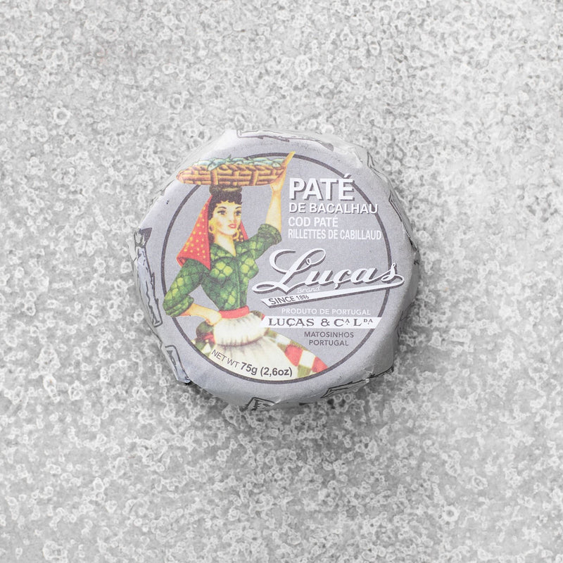 En rund beholder med Lucas Bacalhau torskepaté – portugisisk delikatesse, pakket ind i papir med en traditionel kostumeillustration. Lavet af bæredygtigt fanget torsk, med portugisiske og engelske etiketter.