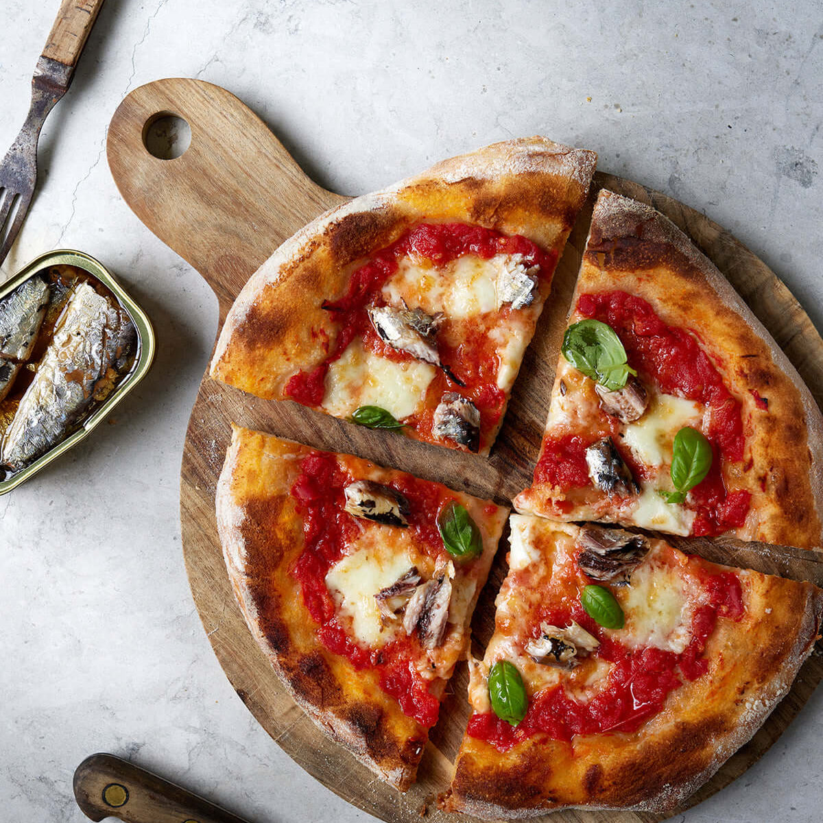 En pizza med tomatsauce, smeltet ost, ansjoser og frisk basilikum er skåret i fire stykker på et træbræt. En dåse sardiner og en gaffel står ved siden af pizzaen på en lys stenoverflade.