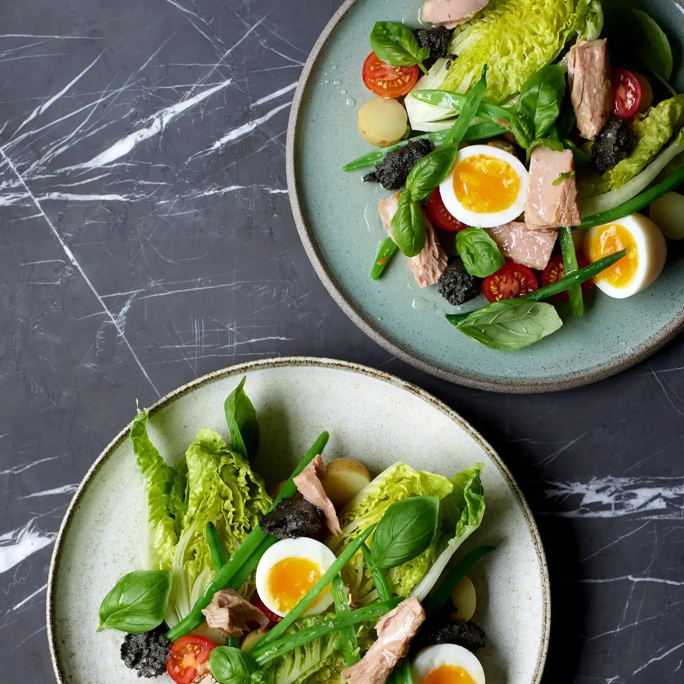 Overblik over to tallerkener med salat, cherrytomater, grønne bønner, kogte æg, kartofler, tun, basilikumblade og klatter af mørk tapenade på en mørk marmoroverflade.