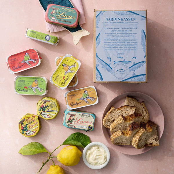 Grøndals Sardinkassen (en æske med assorterede farverige sardinpålæg), skiver brød, superfood-pålæg og to citroner med blade er arrangeret på en lyserød overflade.