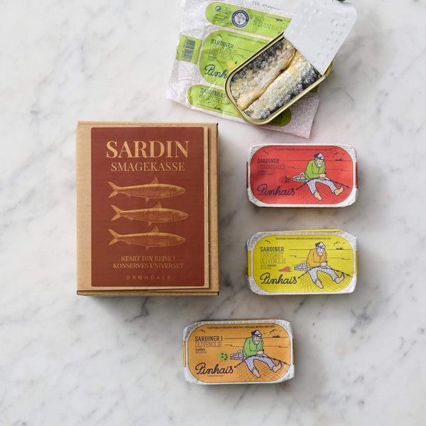 En Grøndals Sardin Smagekasse hviler på marmor ved siden af fire dåser med Portugisiske Sardiner, hvoraf nogle er åbne for at vise fisk, hver i levende rød, gul og blå indpakning.