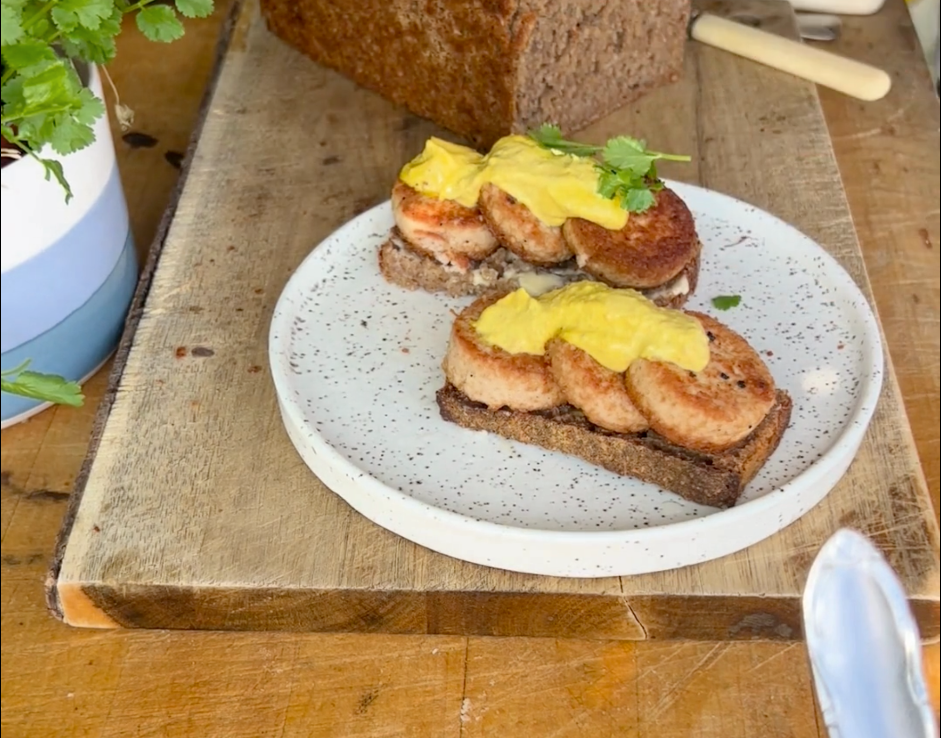 En tallerken med to skiver mørkt brød toppet med brunede pølseskiver, gul sennep og pyntet med friske krydderurter ligger på et træskærebræt ved siden af et brød.