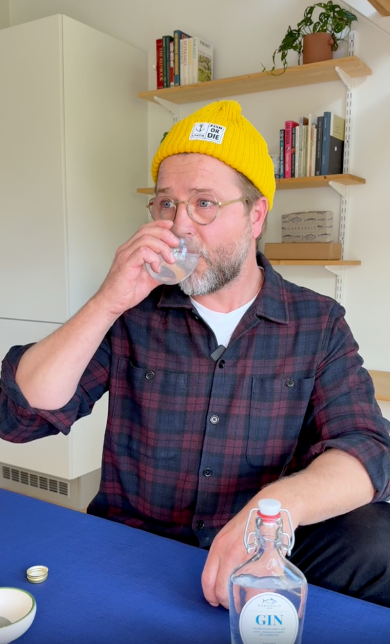 En skægget mand med briller og gul hue drikker af et lille glas, mens han sidder ved et bord med en flaske gin. Bøger og en potteplante står på hylder i baggrunden.