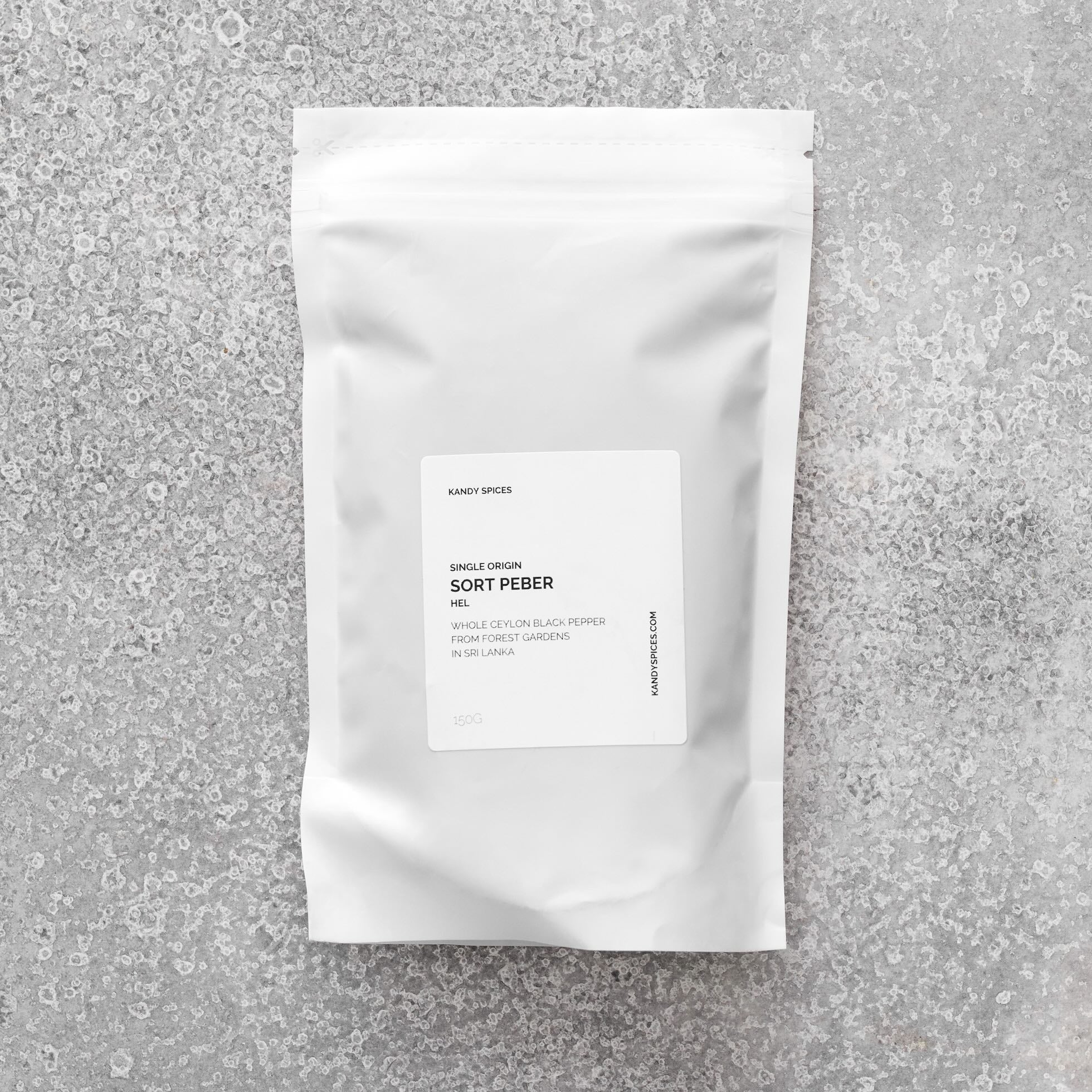 En hvid genlukkelig pose mærket "Sort Peber - i verdensklasse" fra Kandy Spices hviler på en tekstureret grå overflade med et minimalistisk design, der fremhæver dens Ceylon-peber fra Sri Lanka, en enkelt oprindelse.