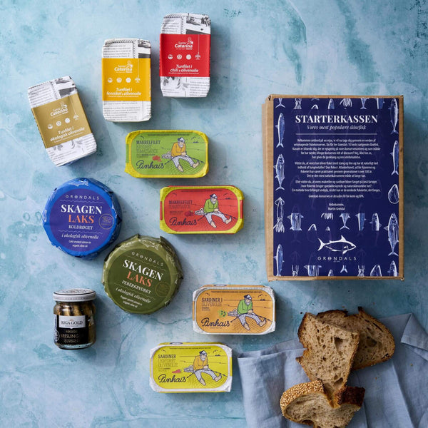 Et kurateret udvalg af Grøndals Starterkassen nordiske gourmet fiskekonserves, paté og pålæg – serveret med rustikt brød og et lille glas – med kvalitetsfisk i BPA-fri dåser på en blå overflade.