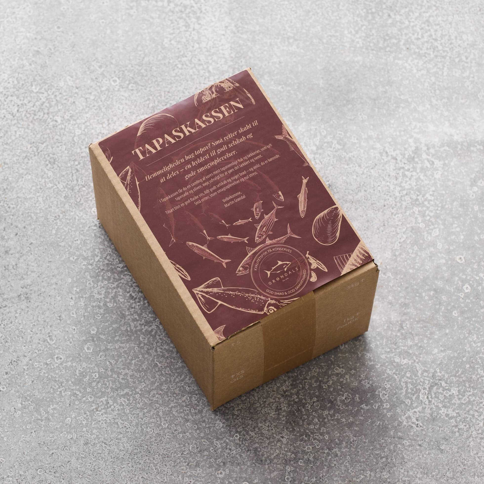 En brun papkasse med en rødbrun TAPASKASSEN-etiket og illustrationer af fisk og køkkenredskaber fra Grøndals indeholder lækre spanske tapas og er udstillet på en grå tekstureret overflade.