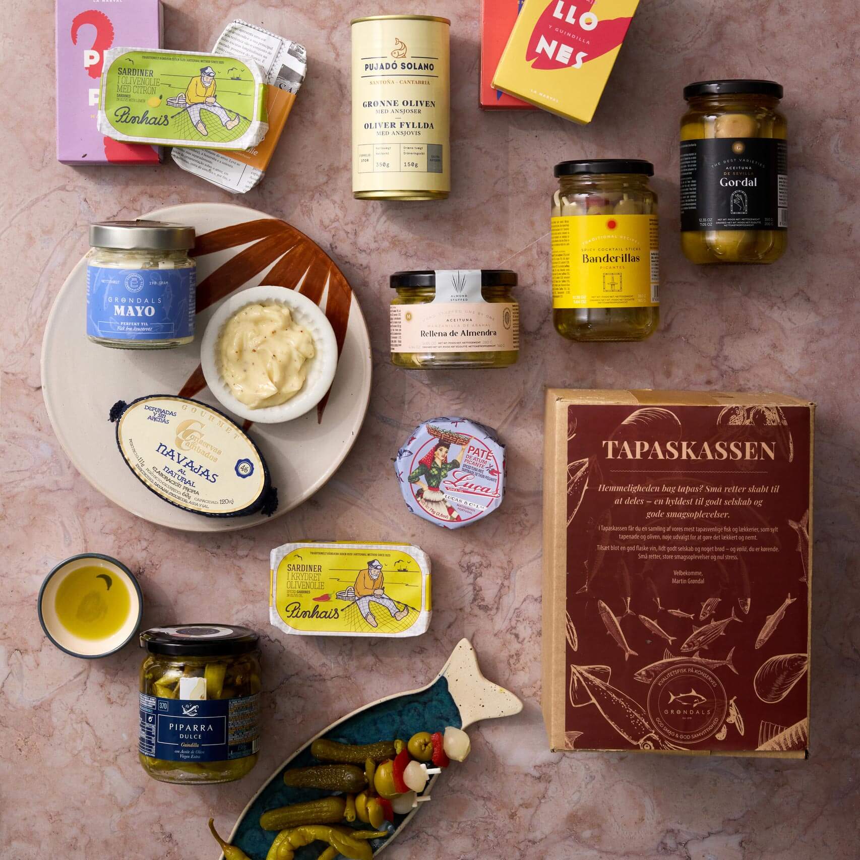 Et udvalg af Grøndals Tapaskassen-tapas med dåseskaldyr, oliven, saucer og syltede grøntsager i farverig emballage på lyserød marmor med Tapaskassen-æsken.