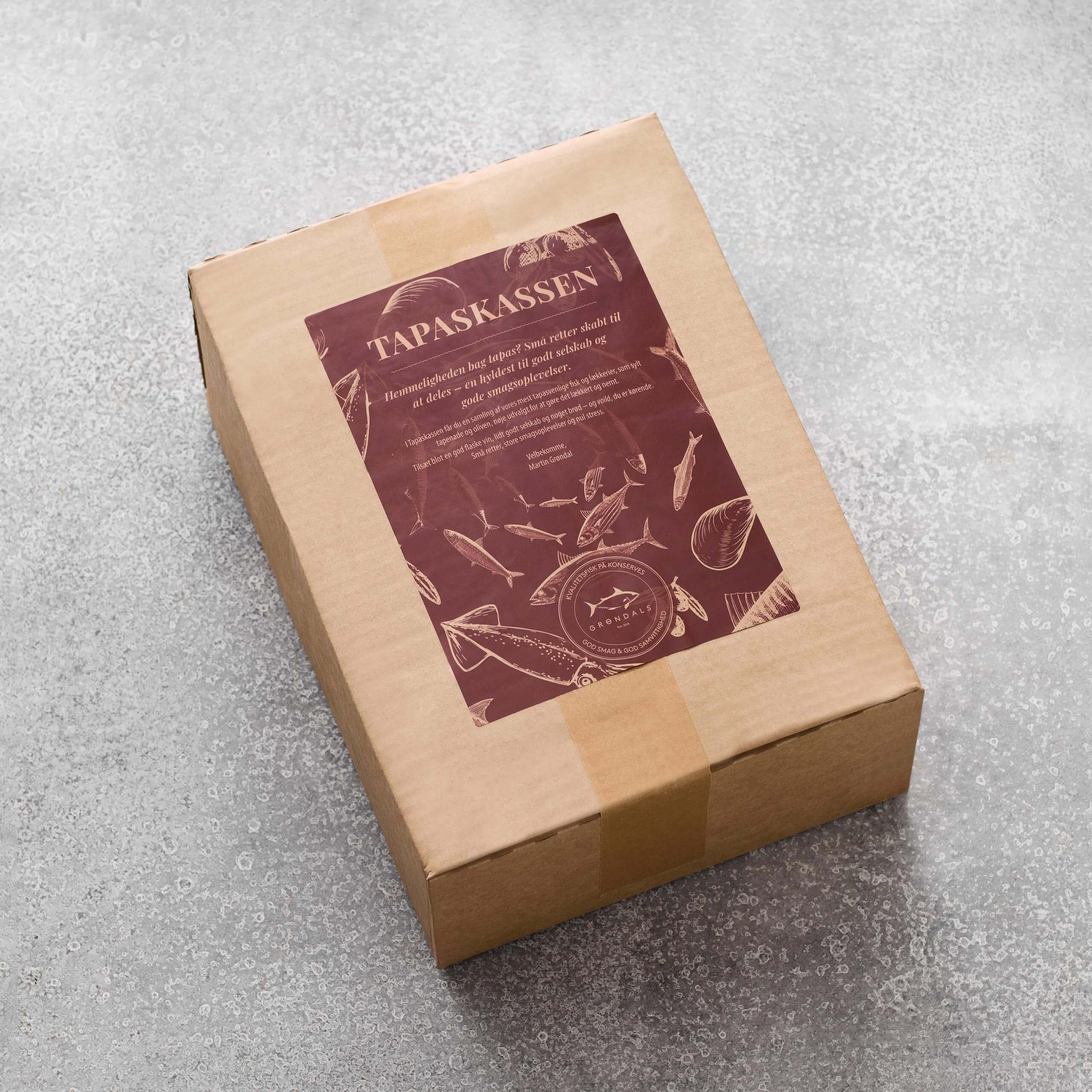 En rektangulær papkasse forseglet med tape, mærket "Grøndals Tapaskassen", med illustrationer af tapasretter som fisk og ost på en lysegrå tekstureret overflade.