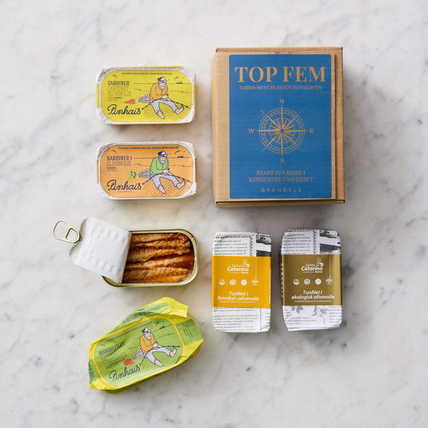 På en marmorplade står dåser med sardiner, en delvist åbnet dåse med synlige fisk, to stablede pakker med gule etiketter og en brun Grøndals "Top Fem Smagekasse" med kompasmotiv.