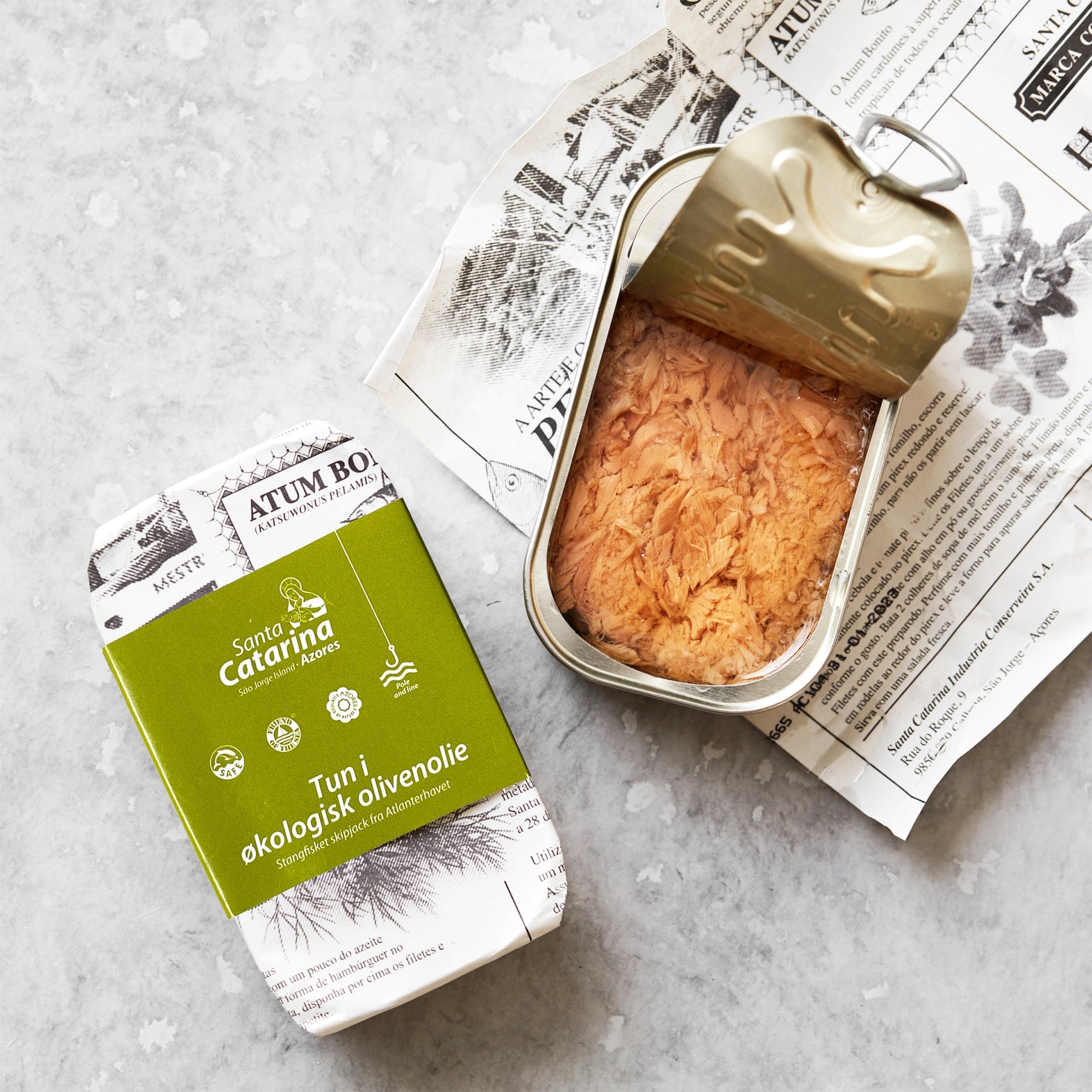 En åbnet dåse med "Småstykker af tun i økologisk olivenolie" fra Santa Catarina ligger ved siden af emballagen og et stykke trykt papir på en lys overflade. Emballagen fremhæver bæredygtig tun i BPA-fri dåser.