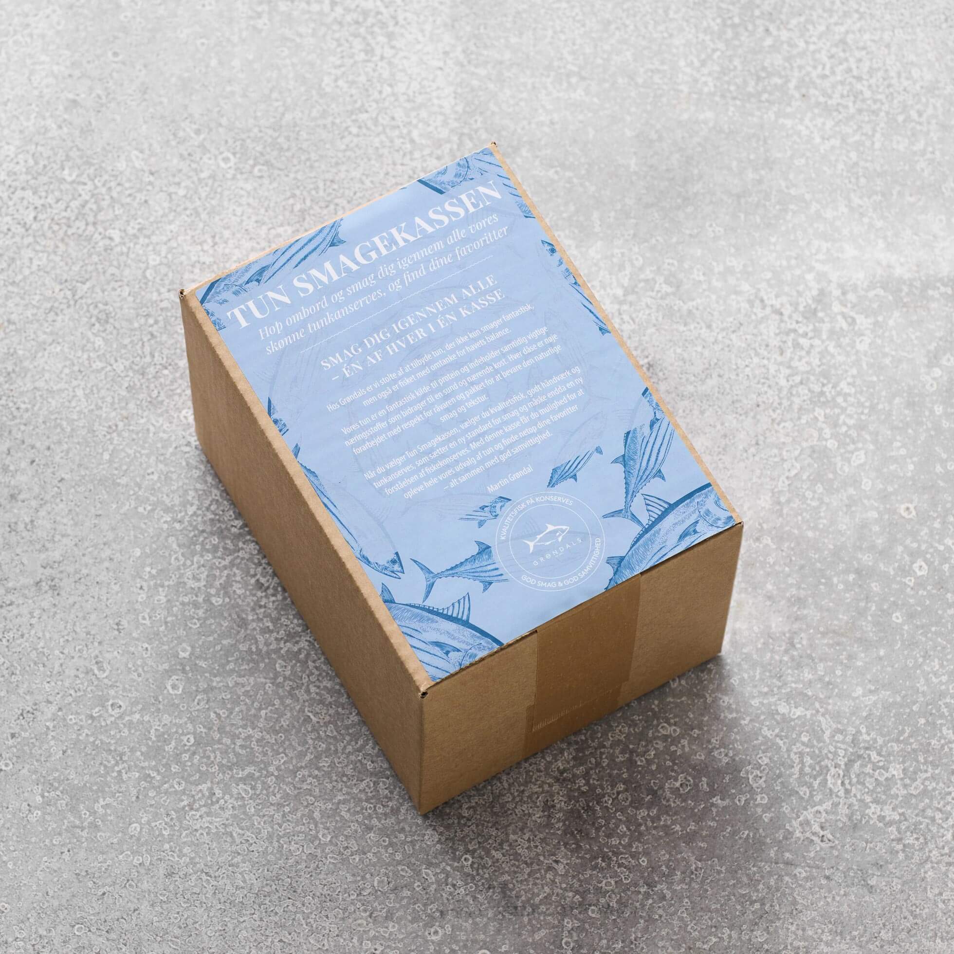 En brun papkasse med en blå etiket påskriften "Tun Smagekassen" fra Grøndals, prydet med hvid tekst og bladillustrationer, står på en grå tekstureret overflade – indeni er der lækkerier som dåsetun i økologisk olivenolie.
