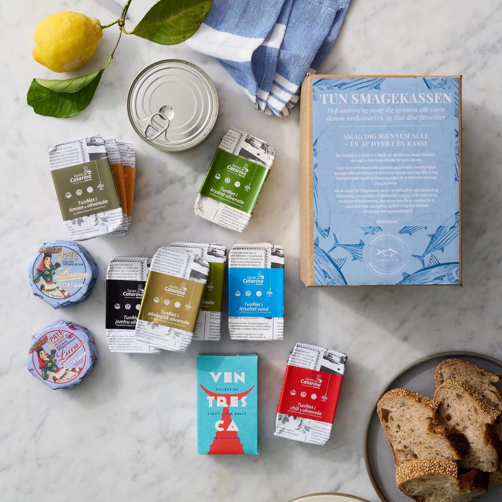 En marmoroverflade viser Grøndals’ Tun Smagekassen-smageæske med Skipjack-tunfilet i økologisk olivenolie, assorteret dåsefisk, paté, brød, en citron og en blå klud.