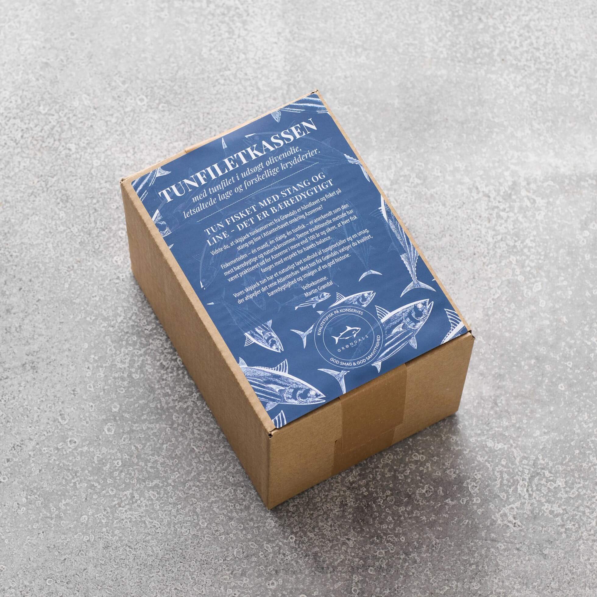 En brun papkasse fra Grøndals ligger på en grå overflade. Den blå etiket med illustrationer af hvid fisk deler detaljer på svensk om Tunfiletkassen – tunfilet i økologisk olivenolie og bæredygtigt fiskeri.