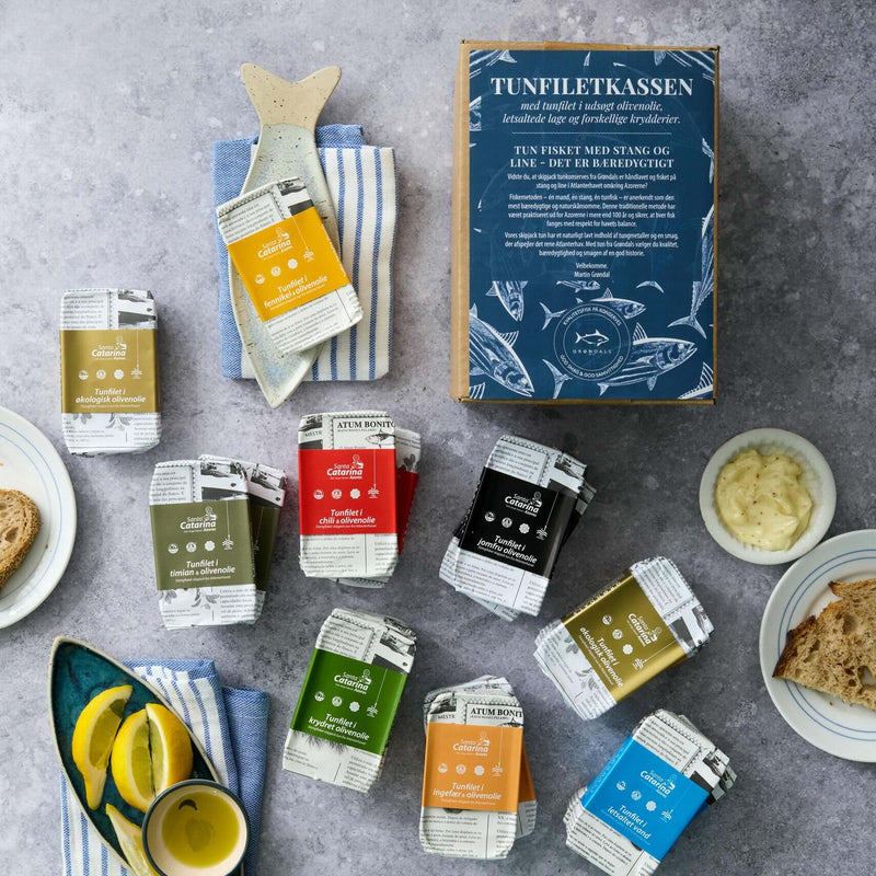 Et udvalg af farverige dåsetun fra Grøndals Tunfiletkassen er udstillet med brød, citronbåde, mayo, stribede klude og en blå æske med påskriften "bæredygtig tun i økologisk olivenolie" på en grå flade.