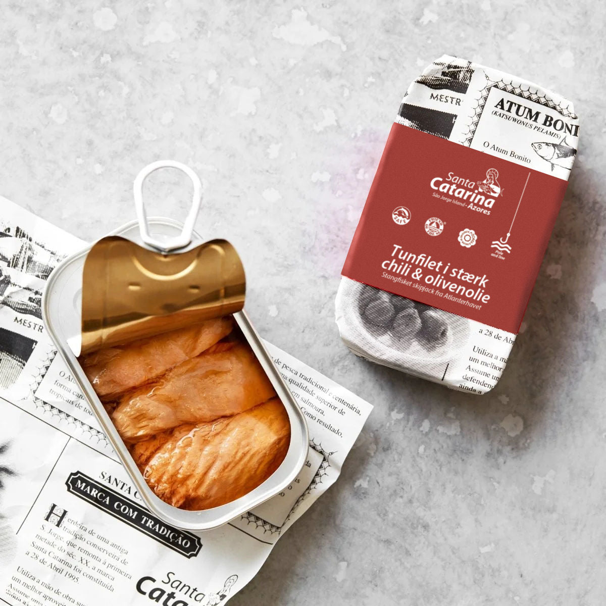 En åben dåse med bæredygtig tun i olie står på et stykke trykt papir ved siden af en lukket rød-hvid dåse med Santa Catarina Tunfilet i stærk chili og olivenolie på en lysegrå overflade.