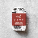 En dåse Santa Catarina Tunfilet med stærk chili og olivenolie, delvist indpakket i papir med tekst og billeder, ligger på en lysegrå, struktureret overflade.