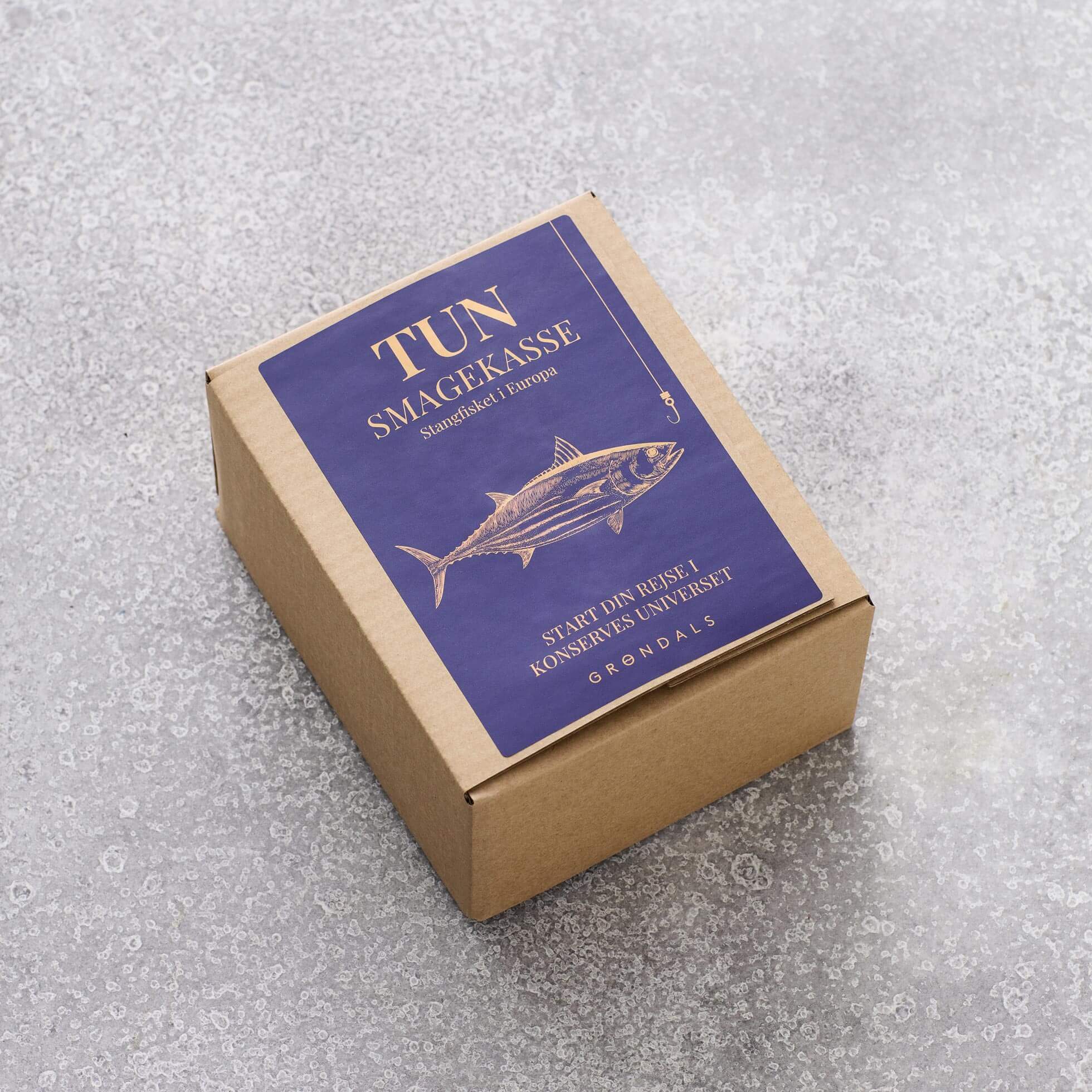 En papæske med en blå etiket med en tunillustration og dansk tekst fra Grøndals, placeret på en lysegrå overflade - ideel til at præsentere bæredygtig tun eller skipjack tun i din Tun Smagekasse.