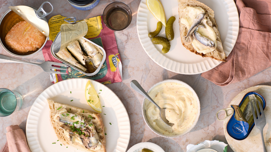 Et billede oppefra og ned af tallerkener med brød med sardiner, syltede agurker, en skål med mayonnaise, åbnede fiskedåser, citronbåde og glas med vand på et marmorbord med lyserøde servietter.