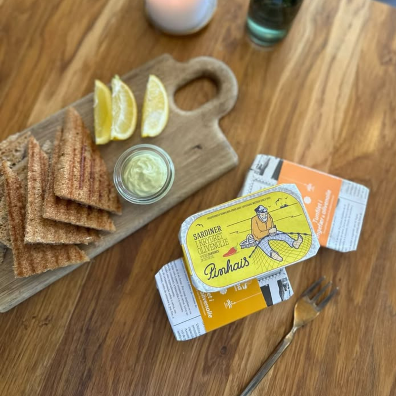 En dåse sardiner i olivenolie står åben på et bord med emballagen ved siden af en træplade med citronbåde, et lille glas sauce og ristede brødskiver. En gaffel ligger i nærheden.
