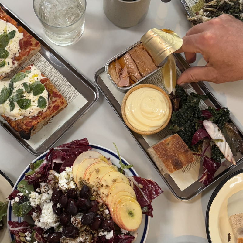En hånd åbner en dåse med fisk ved siden af en bakke med cremet dip, brød og grønt. Der står en salat med æbler, oliven og ost samt rektangulære pizzaslices med krydderurter og et glas isvand på bordet.