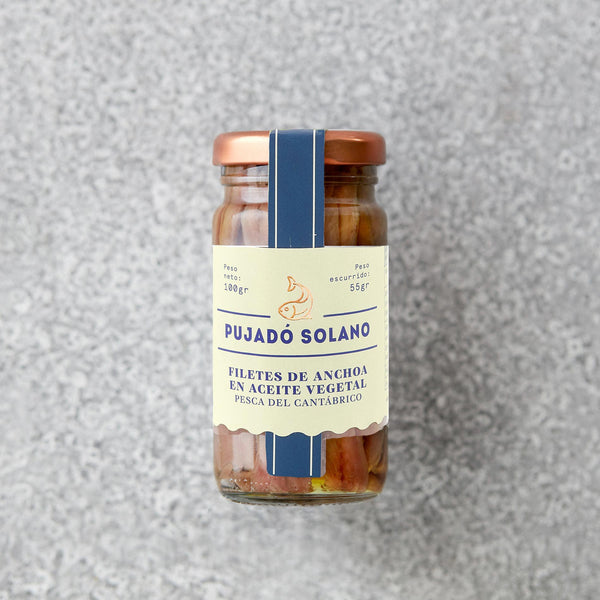 Et sæt på tre Pujado Solano OUTLET saltede ansjosfileter i solsikke, pakket i glaskrukker med beige og blå etiketter med spansk tekst og en fiskeillustration. Placeret på en grå struktureret overflade. Rig på omega-3-fedtsyrer.