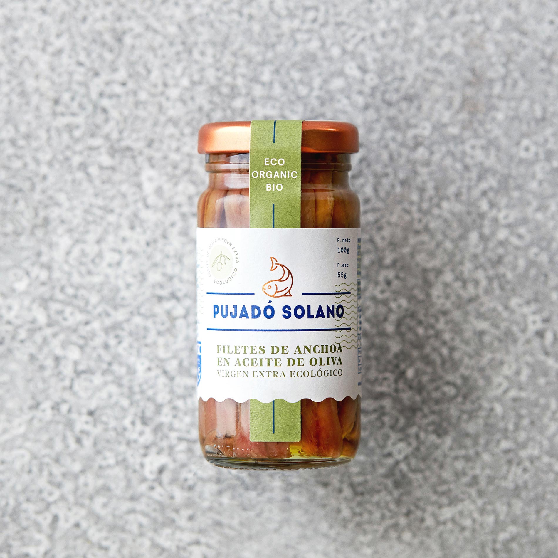En glaskrukke med Pujado Solano OUTLET | Saltede ansjosfileter ØKO olivenolie, med kobberlåg og miljøvenlig grøn etiket, står på en lysegrå struktureret overflade. Rig på omega-3-fedtsyrer.