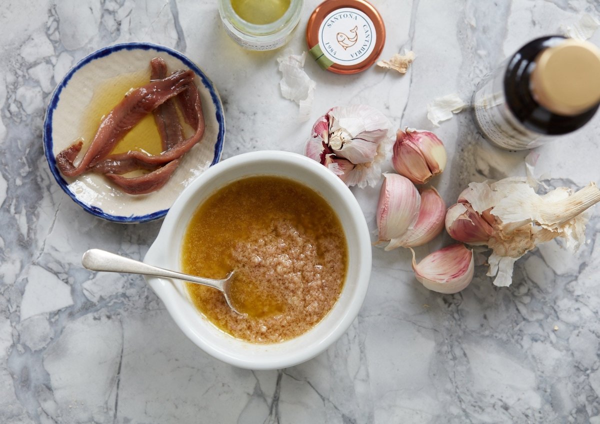 En marmorbordplade med en skål med mosede hvidløg og olie, umami-rige Olasagasti Ansjospuré med olivenolie (60 g), bæredygtige ansjoser, hvidløgsfed, et åbent glaslåg og en flaske olie - alt sammen set fra oven.