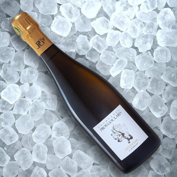 En flaske Champagne Proy-Goulard Éphémère Millésime 2019 BDN med guldfolietop og hvid etiket hviler diagonalt på klare isterninger, og på etiketten står der "Champagne Proy-Goulard.