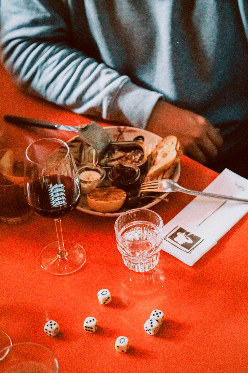 En person sidder ved et rødt bord med en tallerken med forskellige snacks, et glas rødvin, et glas vand, spredte terninger og en serviet med bestik. Kun deres overkrop og arm er synlige.