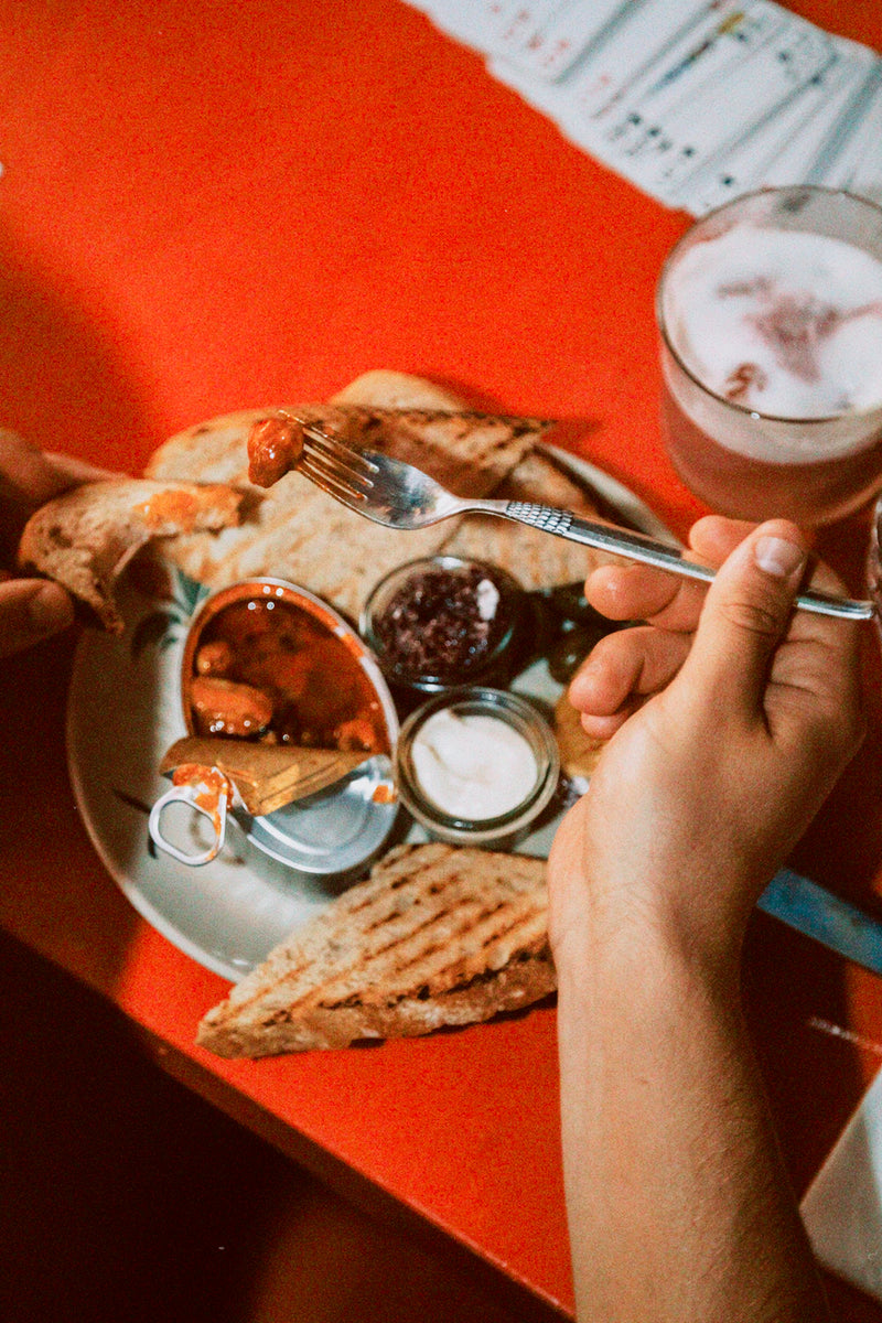 En person holder en gaffel med mad over en tallerken med ristede brødskiver og små krukker med forskelligt pålæg på et rødt bord med en drink og et spil kort i nærheden.