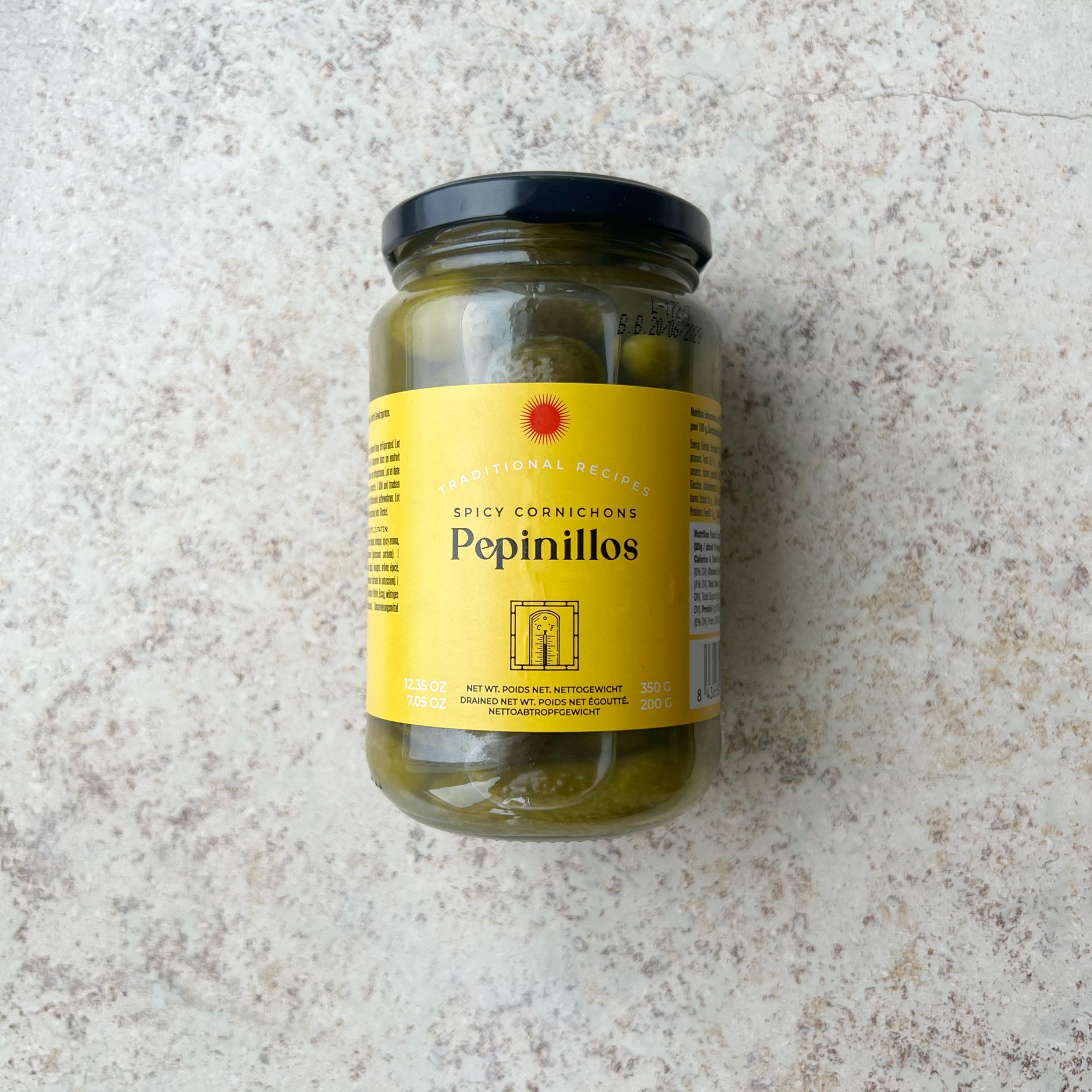 En glaskrukke med Spanske delikatesse cornichoner fra Don Gastronom med en gul etiket og sort låg er præsenteret på en lys, tekstureret overflade; perfekt til servering til tapas.