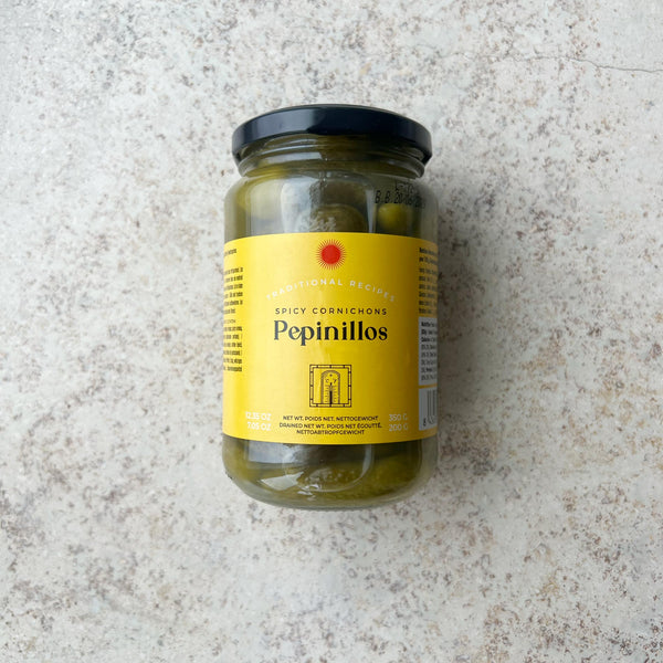 En glaskrukke med Spanske delikatesse cornichoner fra Don Gastronom med en gul etiket og sort låg er præsenteret på en lys, tekstureret overflade; perfekt til servering til tapas.