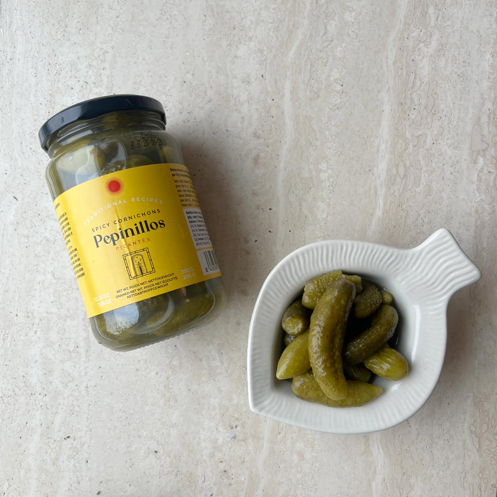 Et glas Don Gastronom Spanske delikatesse cornichoner står på en lys stenoverflade ved siden af en hvid fiskformet skål med syltede agurker, ideelt til din næste tapasaften.