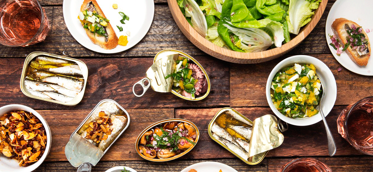 På et rustikt træbord er der arrangeret et udvalg af fisk og skaldyr på dåse, ristet brød med pålæg, frisk grøn salat og glas med rosévin.