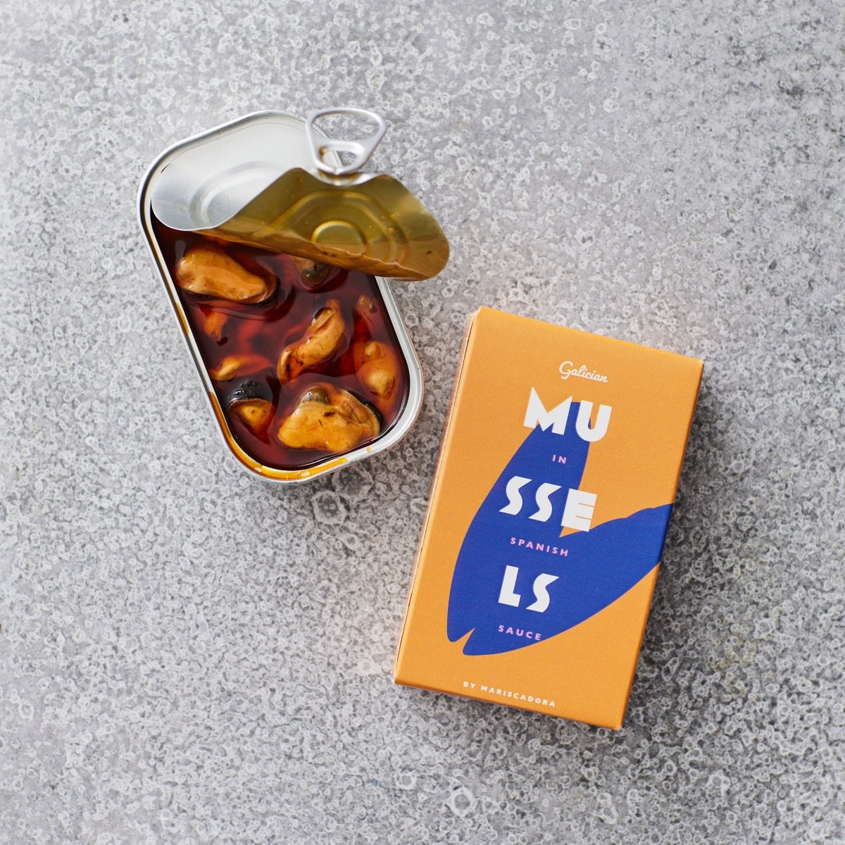 En åben dåse Don Gastronom Friterede muslinger i escabeche ligger ved siden af den orange og blå emballage på en grå, struktureret overflade.