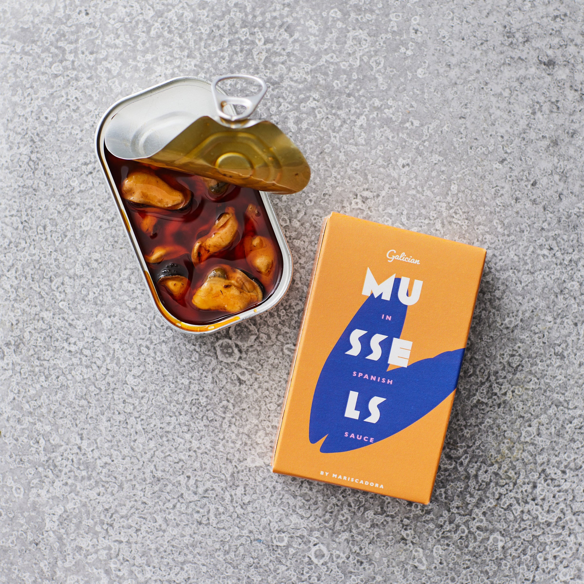 En åben dåse Don Gastronom Friterede muslinger i escabeche ligger ved siden af den orange og blå emballage på en grå, struktureret overflade.