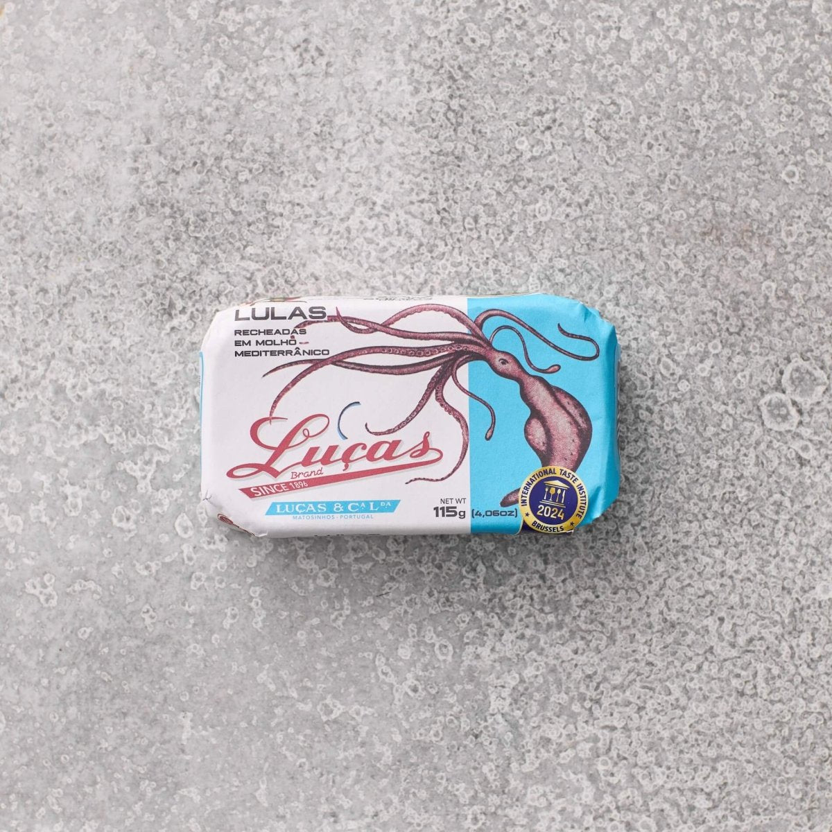 En rektangulær dåse Lucas Fyldte blæksprutte i middelhavssauce med en blæksprutteillustration på den blå og hvide etiket ligger på en tekstureret grå overflade og fremkalder klassiske middelhavsblækspruttesmag.