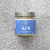 En glaskrukke med Grøndals Mayonnaise – perfekt til fisk fra Grøndals, med et sølvlåg og en blå etiket, hviler på en lysegrå tekstureret overflade. Den danske etiket viser en illustration af en lille fisk, der afspejler mærkets fiskekonserves-arv.
